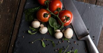 Top view of fresh tomatoes, mushrooms, and greens on a slate board with knife.