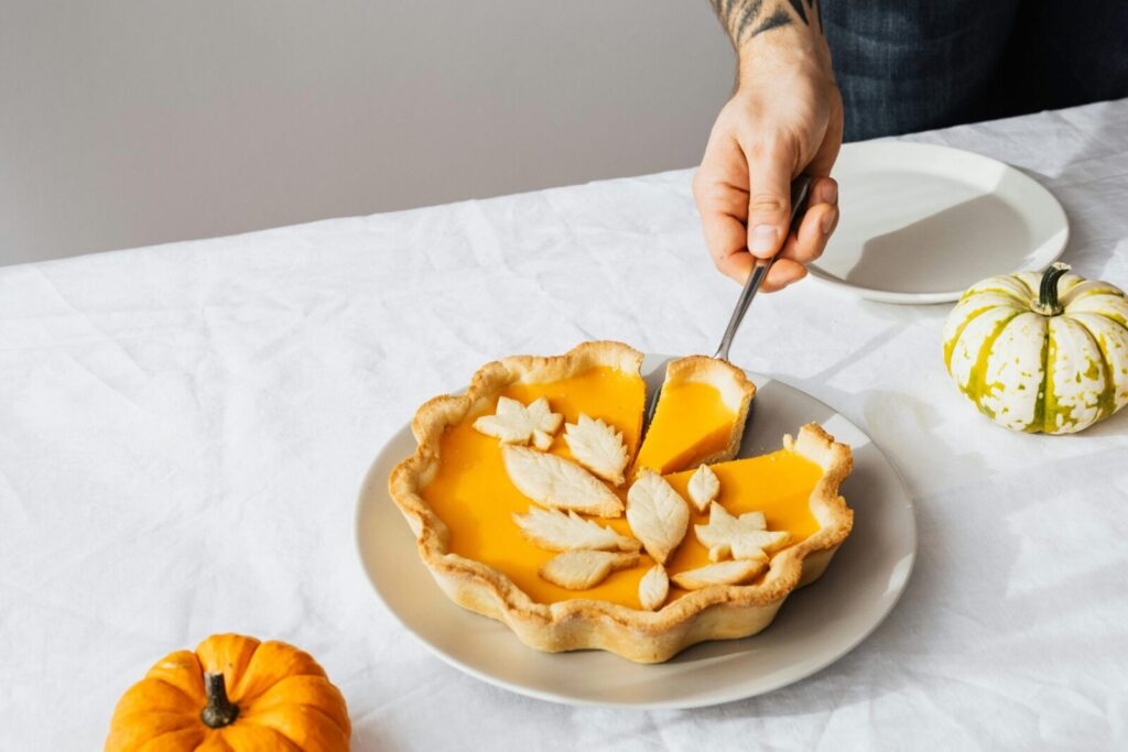 A hand cutting a festive pumpkin pie with decorative crust on a table with small pumpkins.