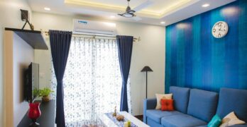 Modern living room with blue accents, featuring a sofa, wall clock, and natural light.