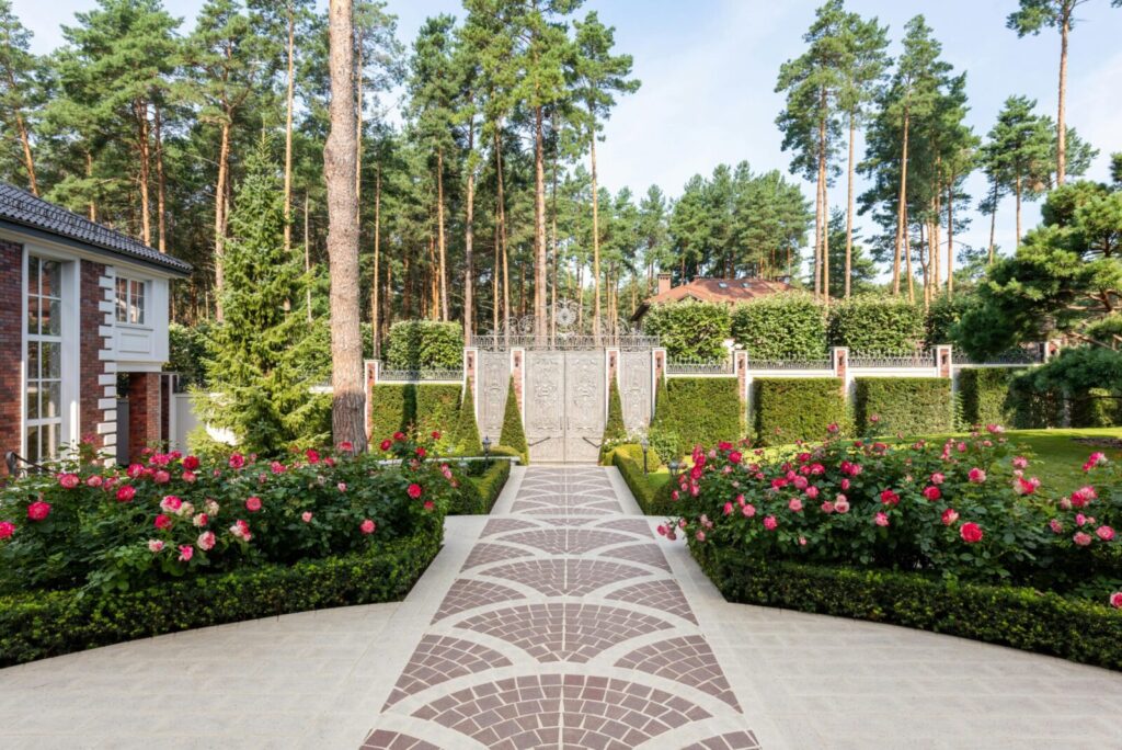 Flowers and green plants with trees in yard with pathways near cottage exterior and fence with gates in sunny summer day