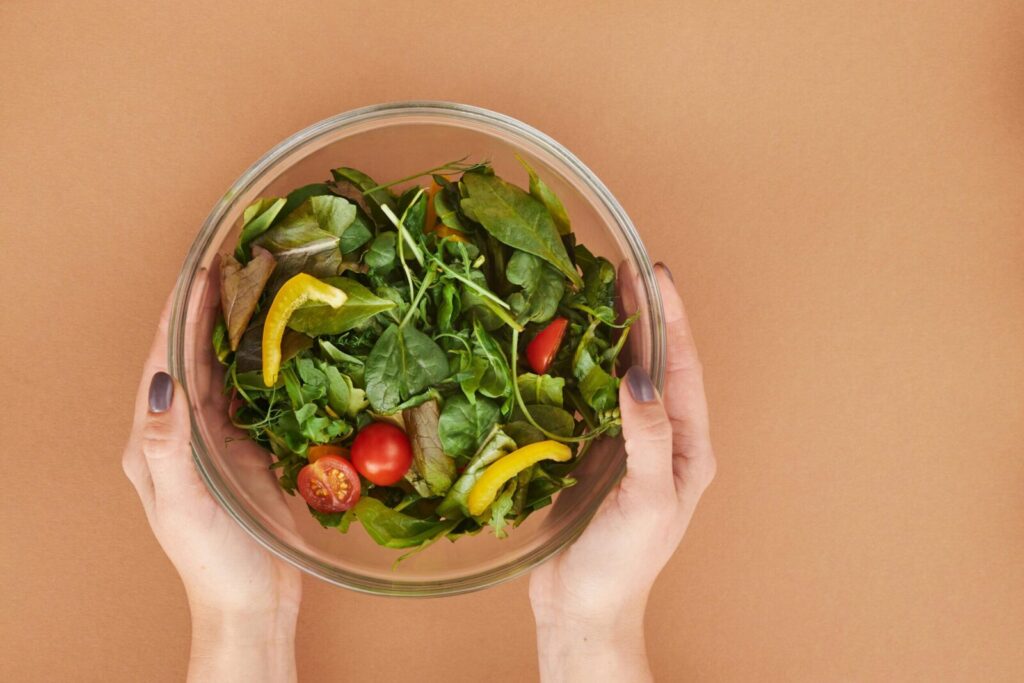 Healthy fresh salad bowl with cherry tomatoes and greens held by hands on a brown surface.