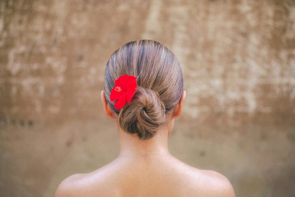 Back view of a woman with a red flower in her hair bun against a rustic backdrop, Bali.