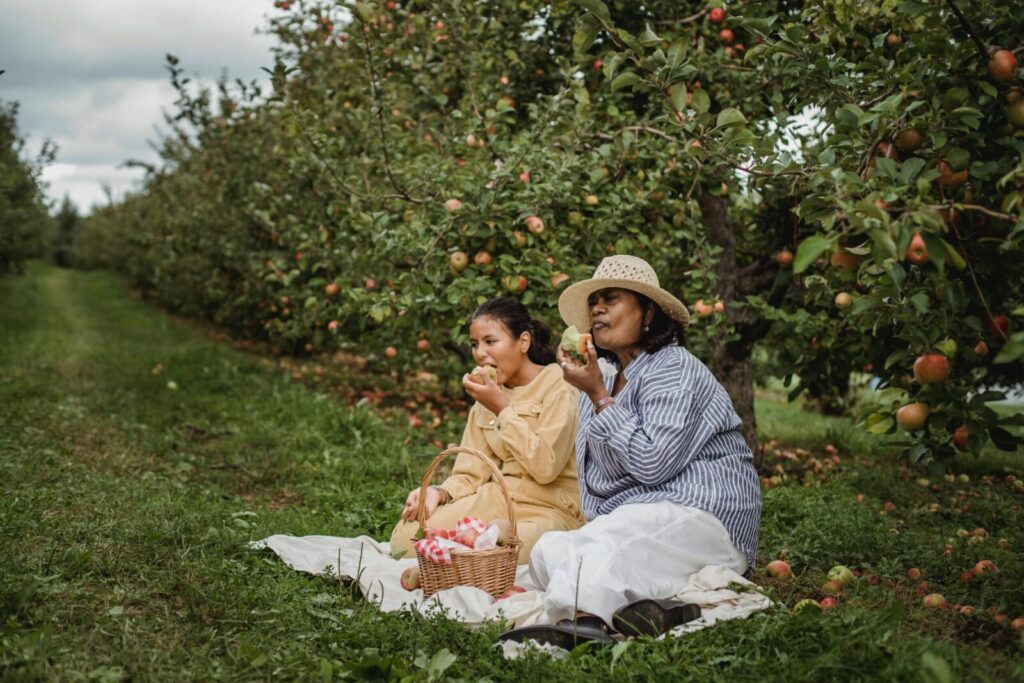 A mother and daughter enjoy a picnic under apple trees, sharing fresh fruit outdoors.