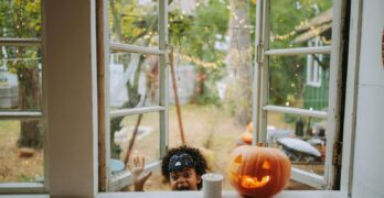 A young black boy celebrates Halloween with a carved pumpkin by an open window.