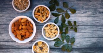 Aerial view of assorted nuts and seeds in white bowls with green leaves on a rustic wooden table.