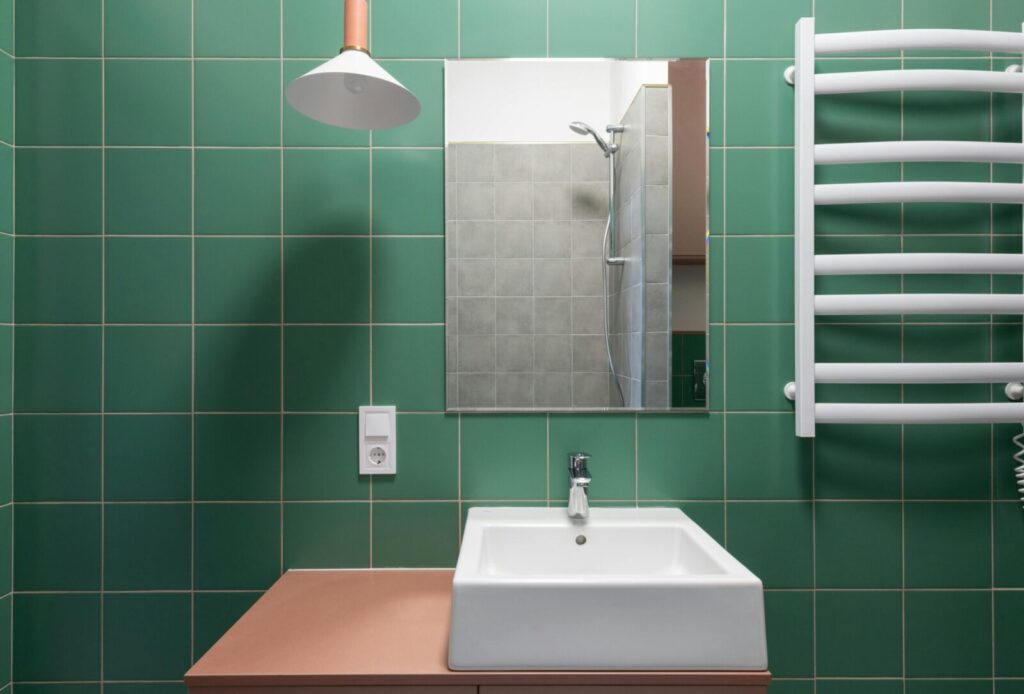 A contemporary bathroom with green tiles, white sink, and mirror reflecting shower.