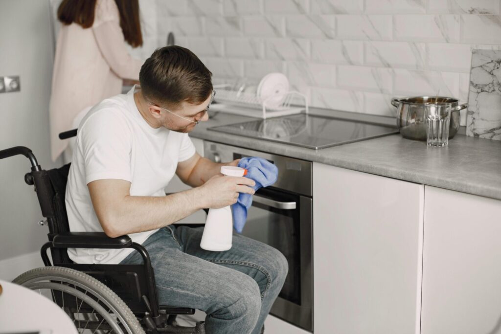 A man using a wheelchair cleans a modern kitchen surface with a spray bottle and cloth.