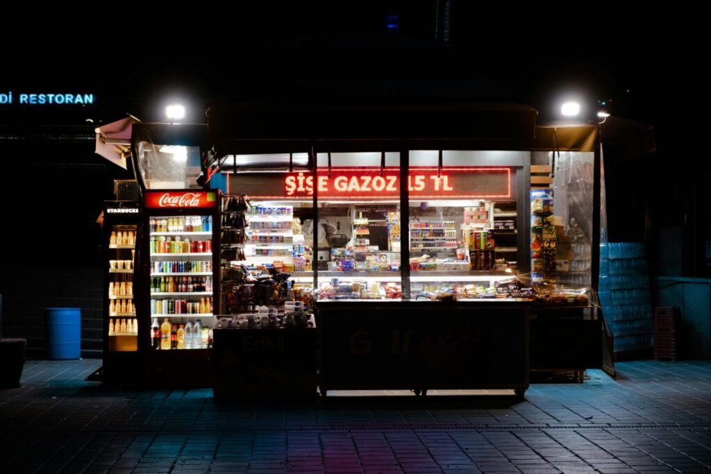 Illuminated nighttime street kiosk with drinks and snacks for sale.