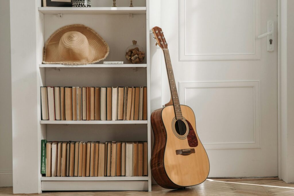 A warm interior scene with an acoustic guitar next to a book-filled shelf and a straw hat.