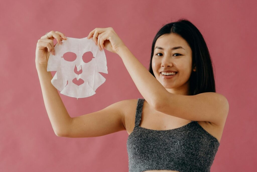 Smiling Asian woman showcasing skincare routine with a sheet mask against a pink backdrop.