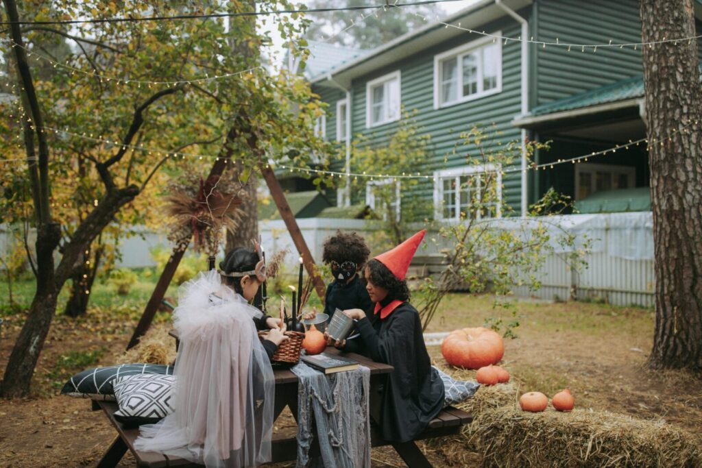 Three children in Halloween costumes sitting at a picnic table with pumpkins in a festive, decorated backyard.
