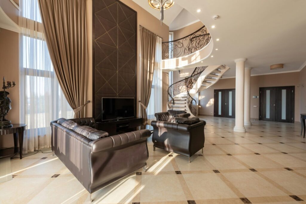 Soft leather black couches on tiled floor near window with curtains in modern stylish house with stairway and doors near columns