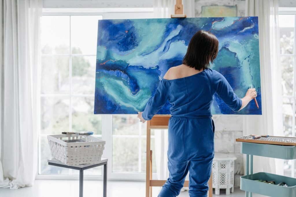 Woman painting an abstract blue design on a large canvas in a bright art studio.