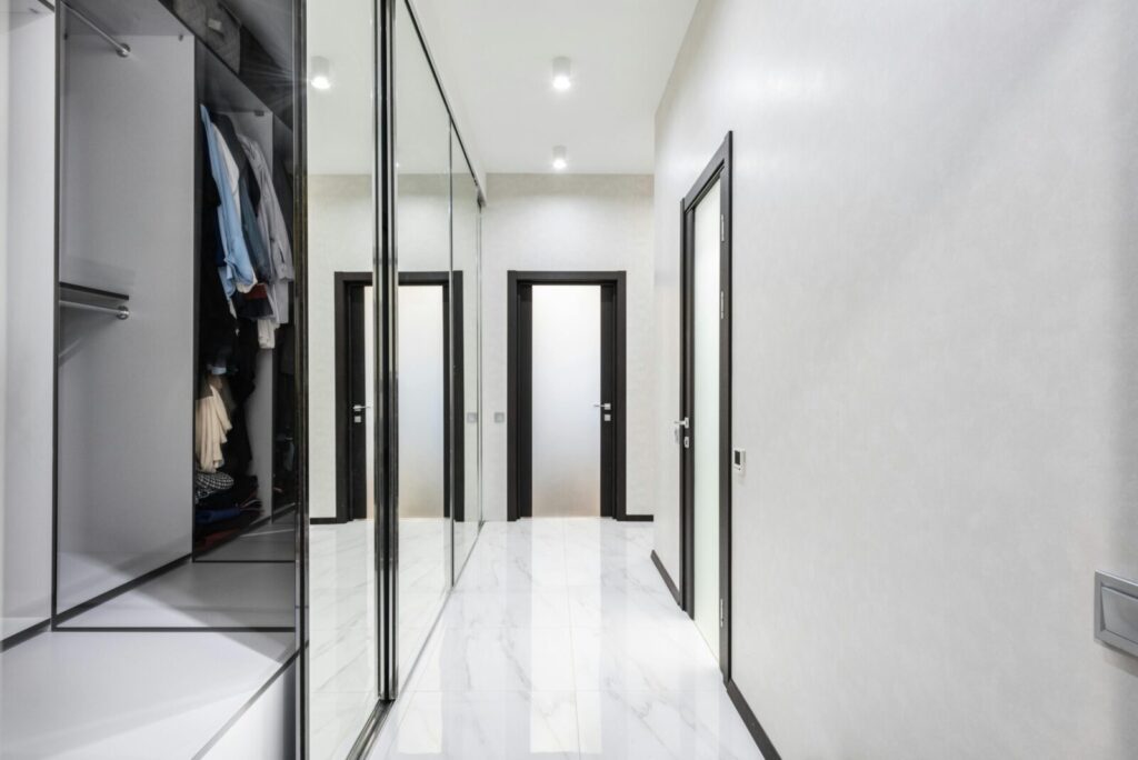 Long light corridor with white walls and opened wardrobe with cloth in modern apartment with closed doors and glowing lamps