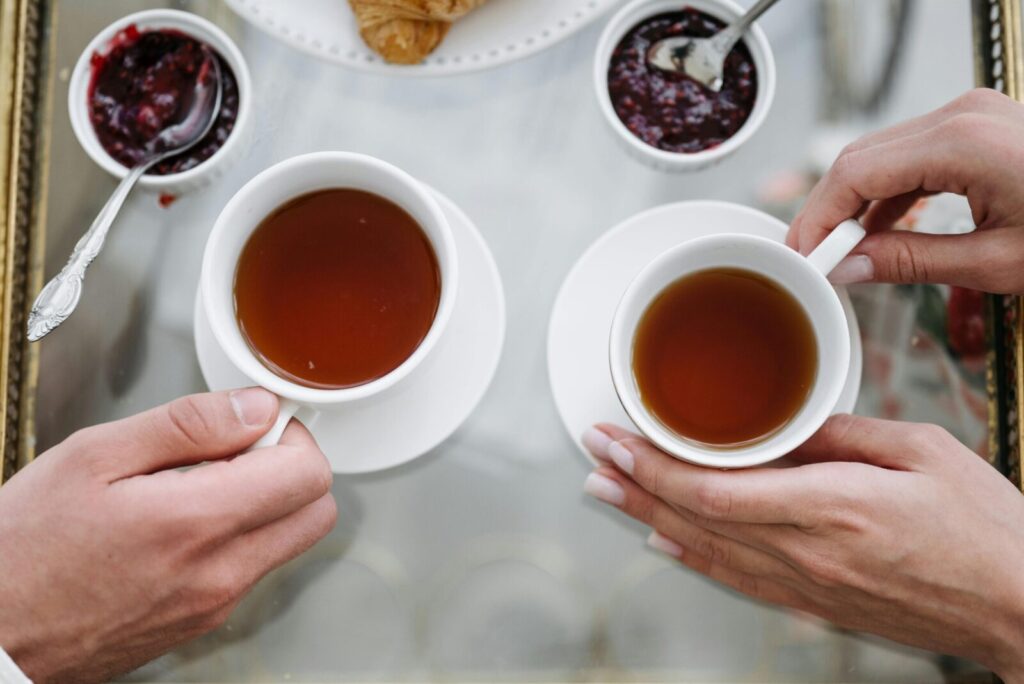 Top view of two cups of tea with hands, jam, and croissants, creating a cozy atmosphere.