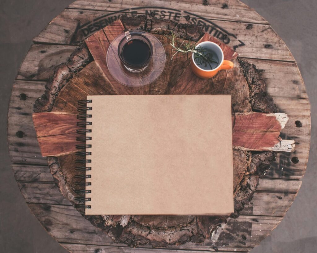 Top view of a rustic table with a blank notebook, coffee, and herbal tea.