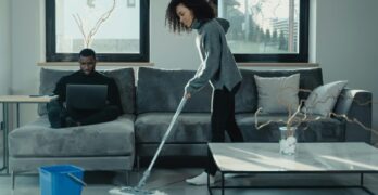 Man working on laptop while woman mops floor in stylish living room.