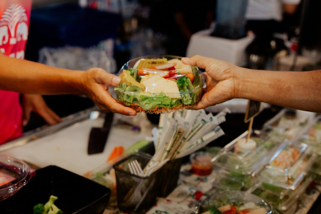 A person hands over a fresh salad in a plastic container at a food stall.
