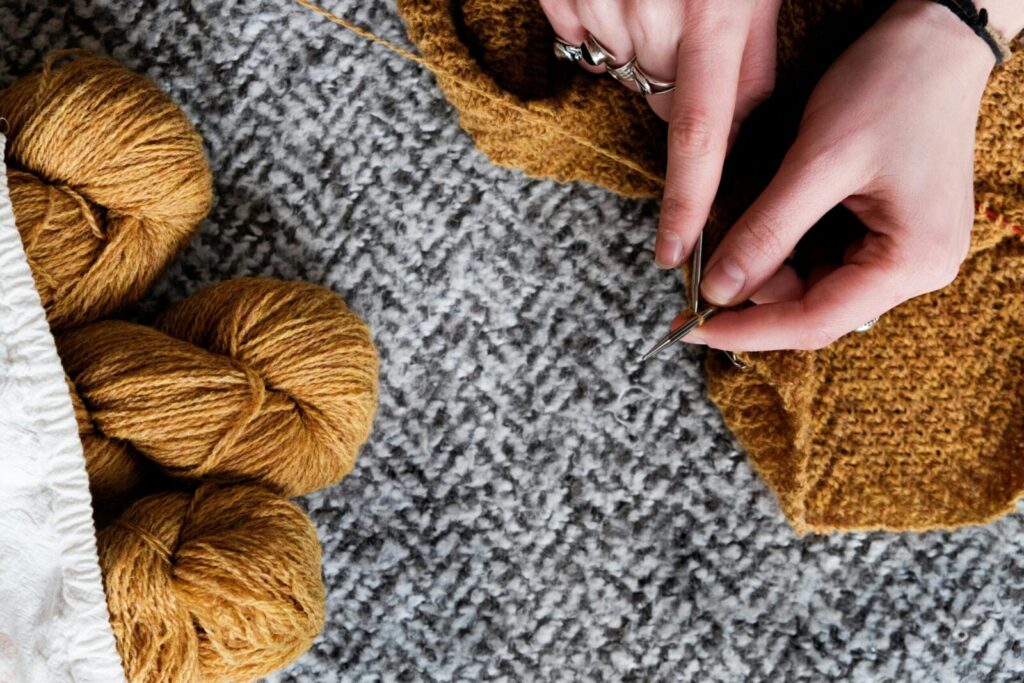 Close-up of hands knitting mustard wool with a gray textured carpet background.