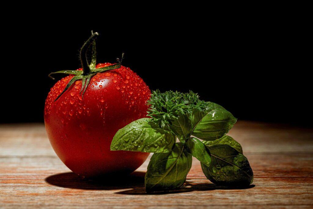 Vibrant red tomato and green basil leaves with water droplets on a wooden surface.