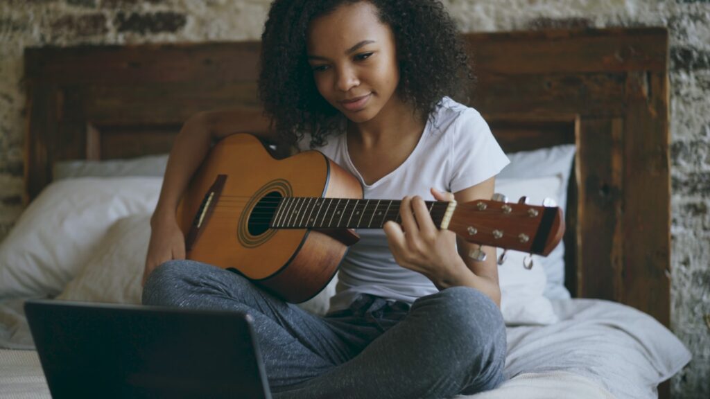 Young woman learning guitar from laptop on bed