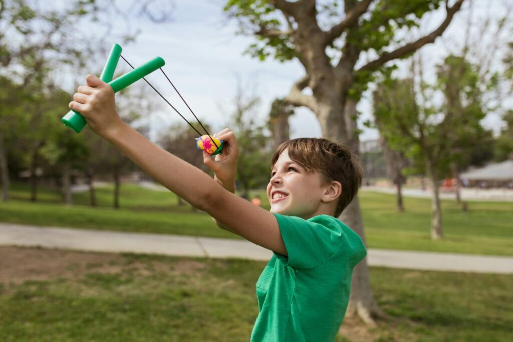 A cheerful boy playing with a slingshot in a sunny park setting.