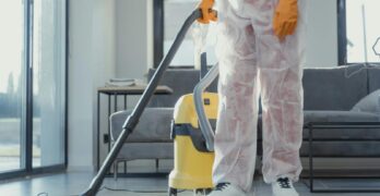 A cleaner in protective coveralls and gloves vacuuming a modern living room.