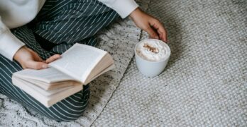 From above of crop anonymous female sitting with crossed legs and reading book with mug of coffee with whipped cream while resting at home