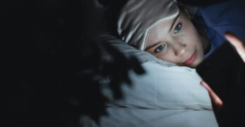 A woman in bed wearing a sleep mask uses her phone at night, creating a cozy ambiance.