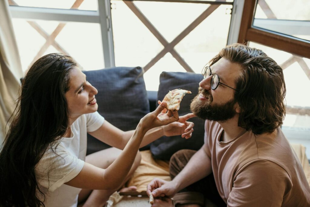 A bearded man and woman share a slice of pizza indoors, enjoying a relaxed moment.