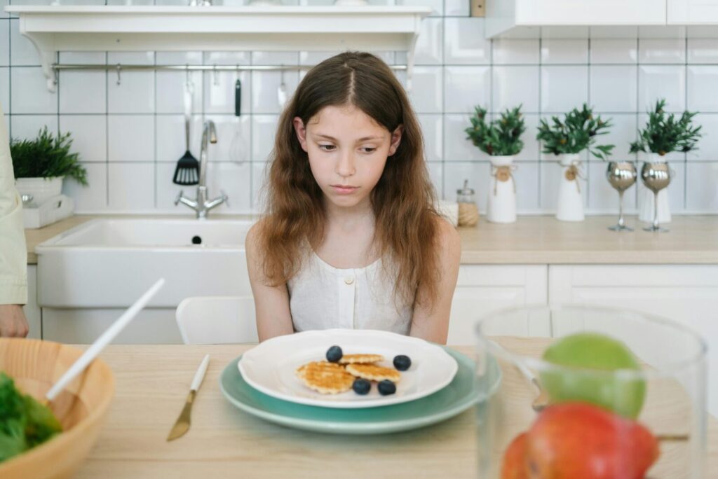A young girl looks down unhappily at her breakfast pancakes with blueberries in a modern kitchen.