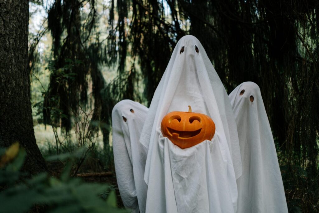 Three ghostly figures holding a carved pumpkin in a mysterious forest setting, perfect for Halloween imagery.