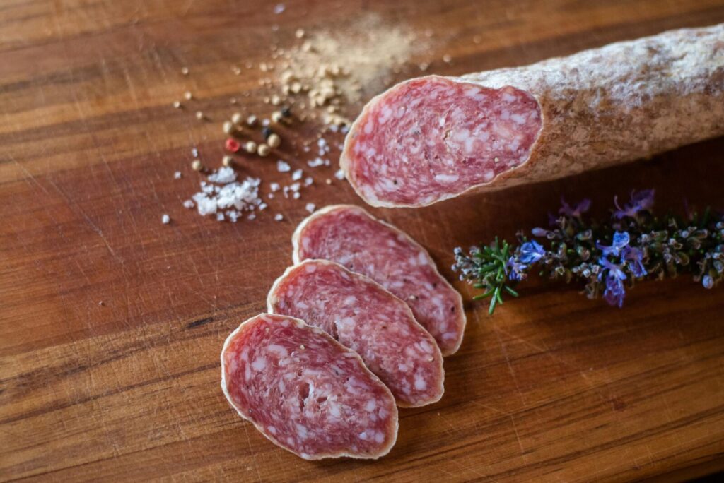 Tasty sliced salami on wooden board with rosemary and assorted peppercorns.