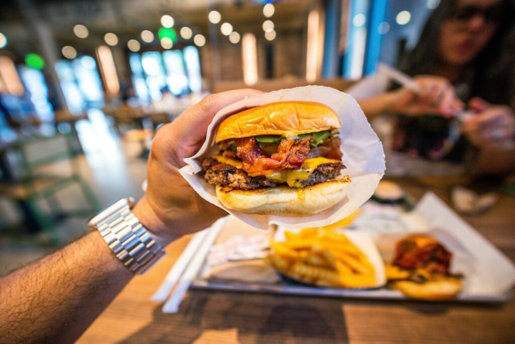 Close-up of a tasty bacon cheeseburger held in hand, with fries in a casual restaurant setting.