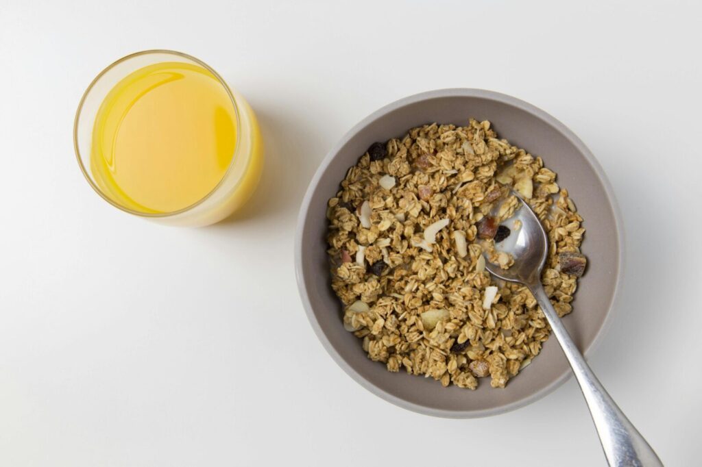 A top view of granola in a bowl with a spoon and a glass of orange juice on a white background.