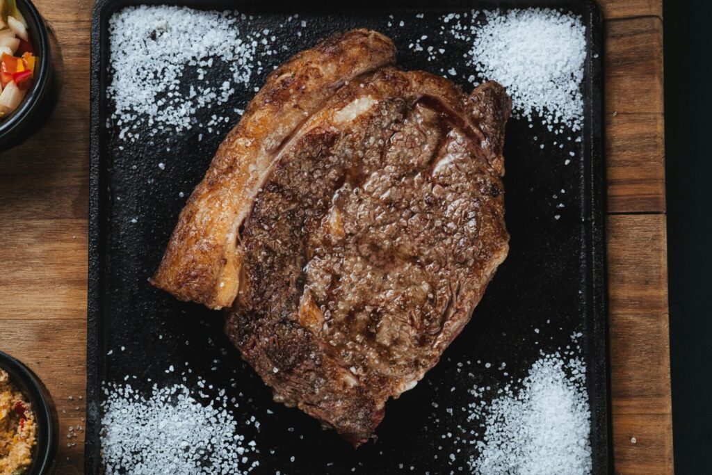 Grilled ribeye steak on a black surface with salt, showcasing juicy meat texture.
