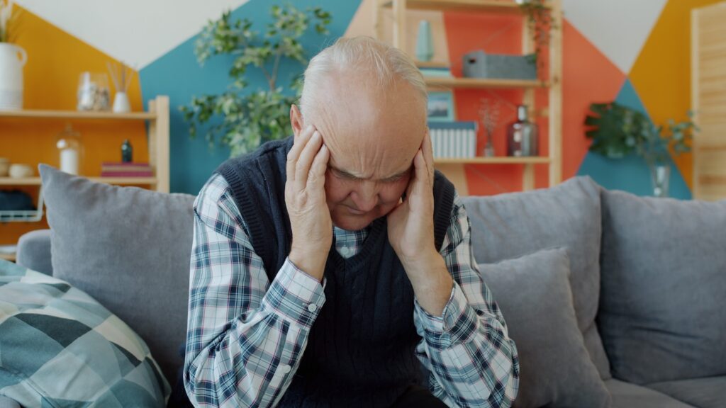 Elderly man holding head in discomfort on couch
