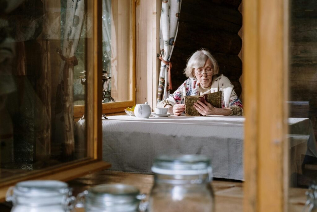 Senior woman reads by the window in a cozy cabin, enjoying a cup of tea with a teapot on the table.