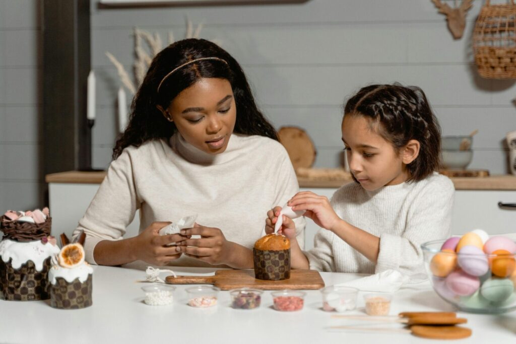 African American woman and girl decorating cupcakes with icing and candy at home.