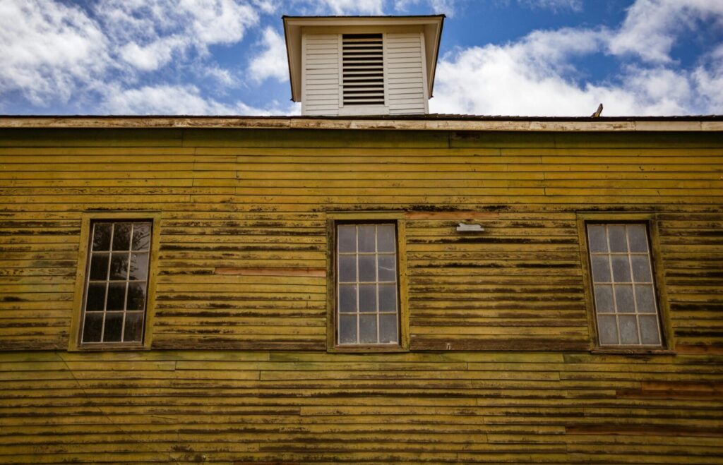 A rustic vintage building with wooden siding and three windows against a blue sky.