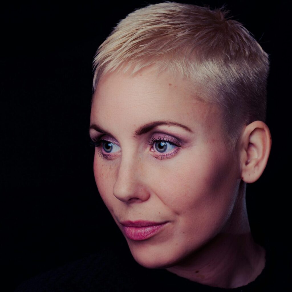 Close-up portrait of a woman with short blonde hair against a dark background.