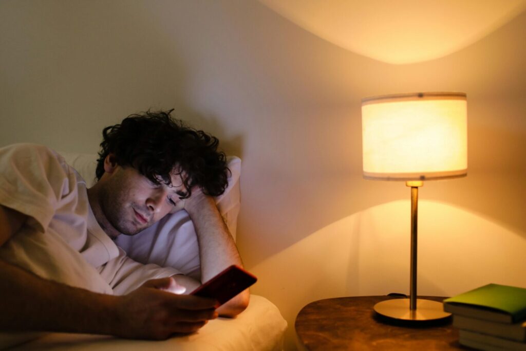 A man lying in bed using a smartphone with a lamp providing warm ambient light.