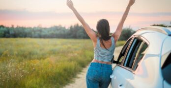 A woman stands beside a car on a summer field road, arms raised in freedom, enjoying a beautiful sunset.