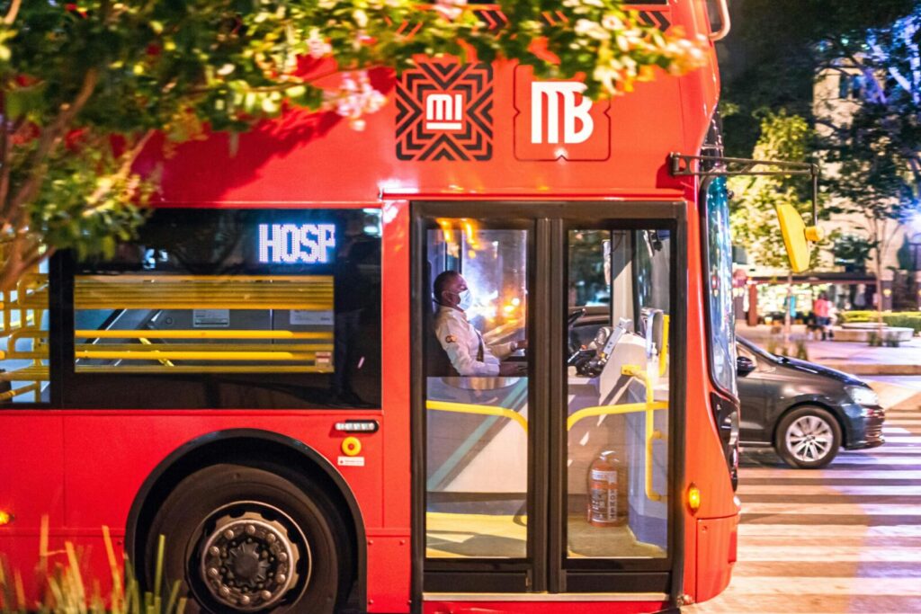 Red double-decker bus with masked driver on a bustling evening street in Ciudad de México.