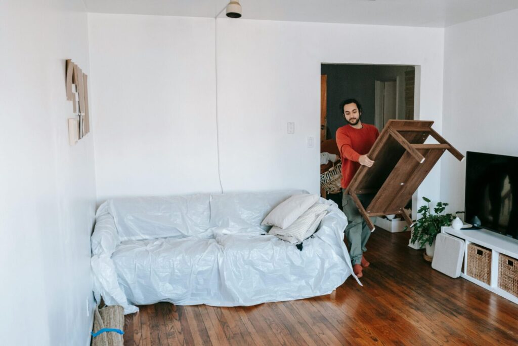Man lifting a wooden table in a bright living room with wrapped sofa, preparing for a move.