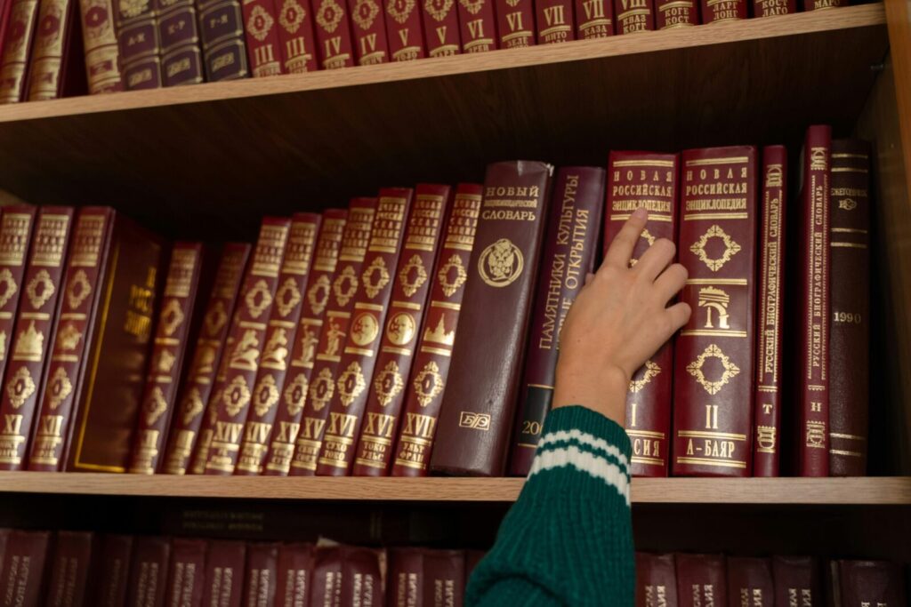 A person reaching for a red encyclopedia on a neatly organized bookshelf in a library.