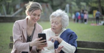 Delighted female relatives sitting together on wooden bench in park and browsing mobile phone while learning using