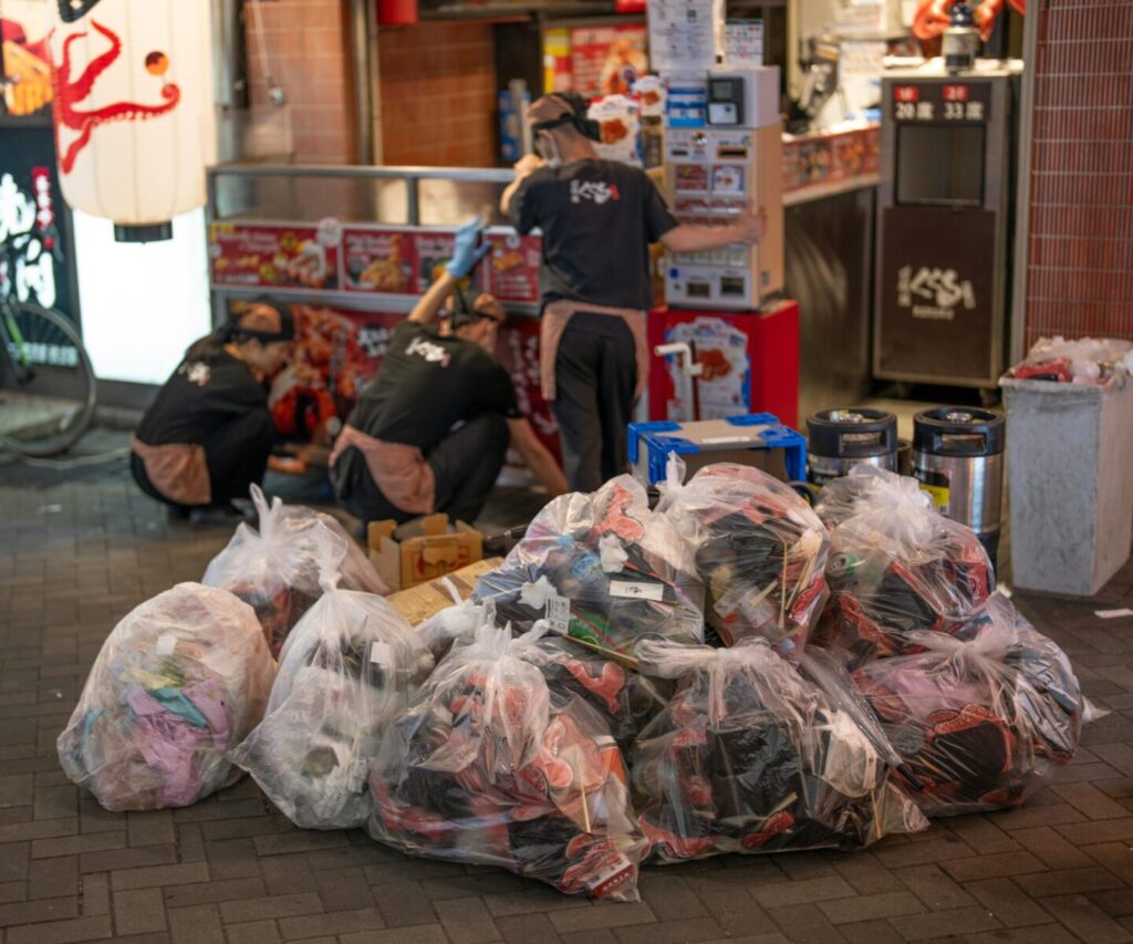 Workers tidying a Japanese street market with waste bags. Urban commerce scene.