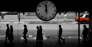 People walk by a clock on a busy street.
