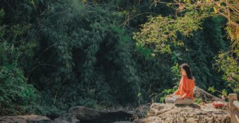 Woman meditating by a serene riverside surrounded by lush greenery on a sunny day.